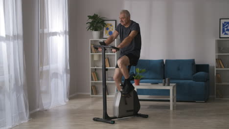 healthy-lifestyle-in-middle-age-man-is-training-on-stationary-bicycle-in-room-keeping-good-physical-condition-wide-shot